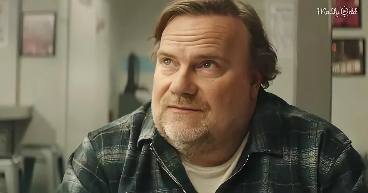A man with a beard and plaid shirt looks off to the side in a room with posters on the wall.