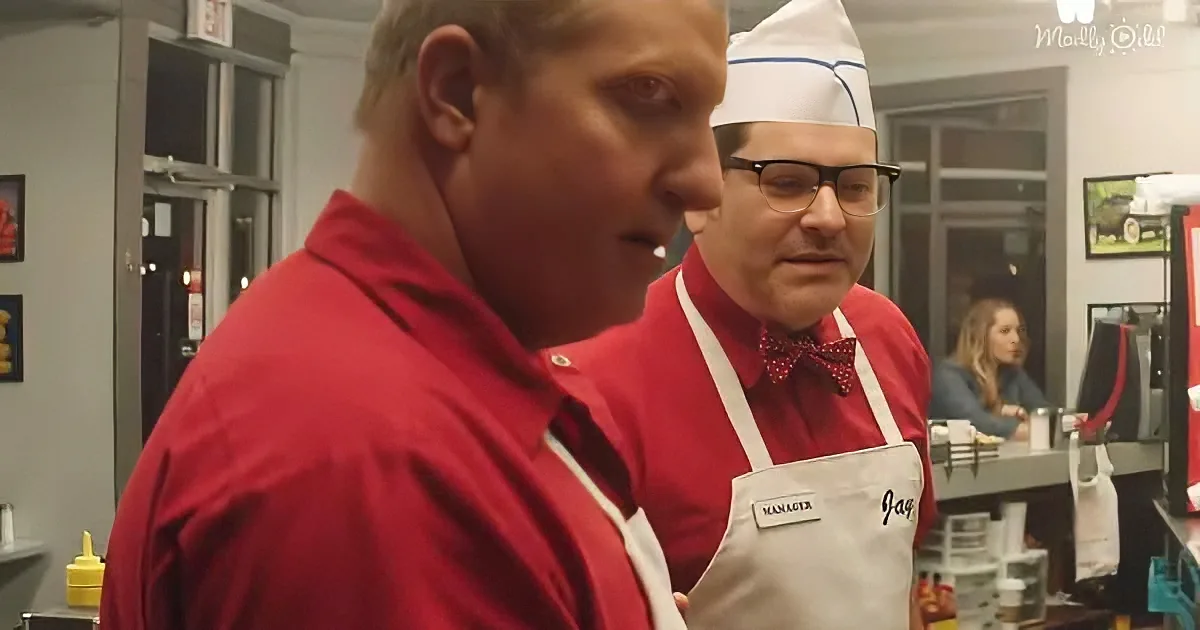 A man in a red shirt and chef's hat stands next to another man in a red apron and bow tie, both in a kitchen setting.