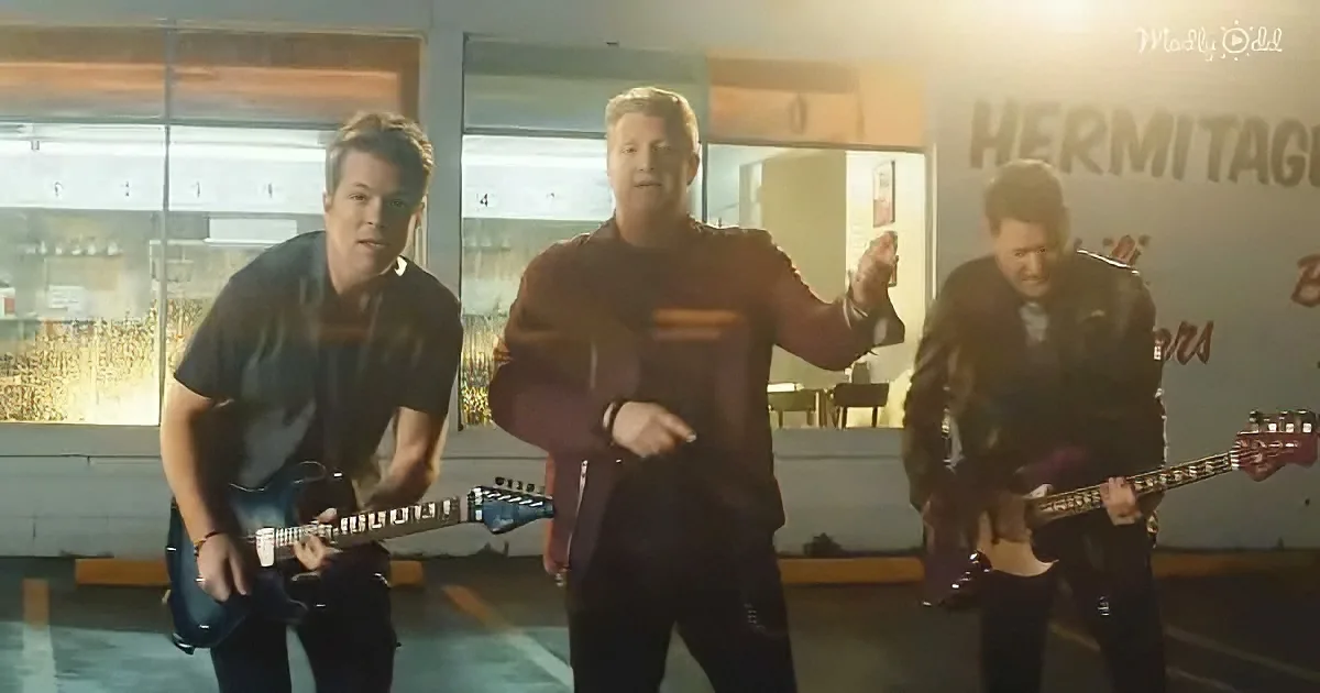 A man in a red shirt dances energetically while two others play guitars in a brightly lit room.