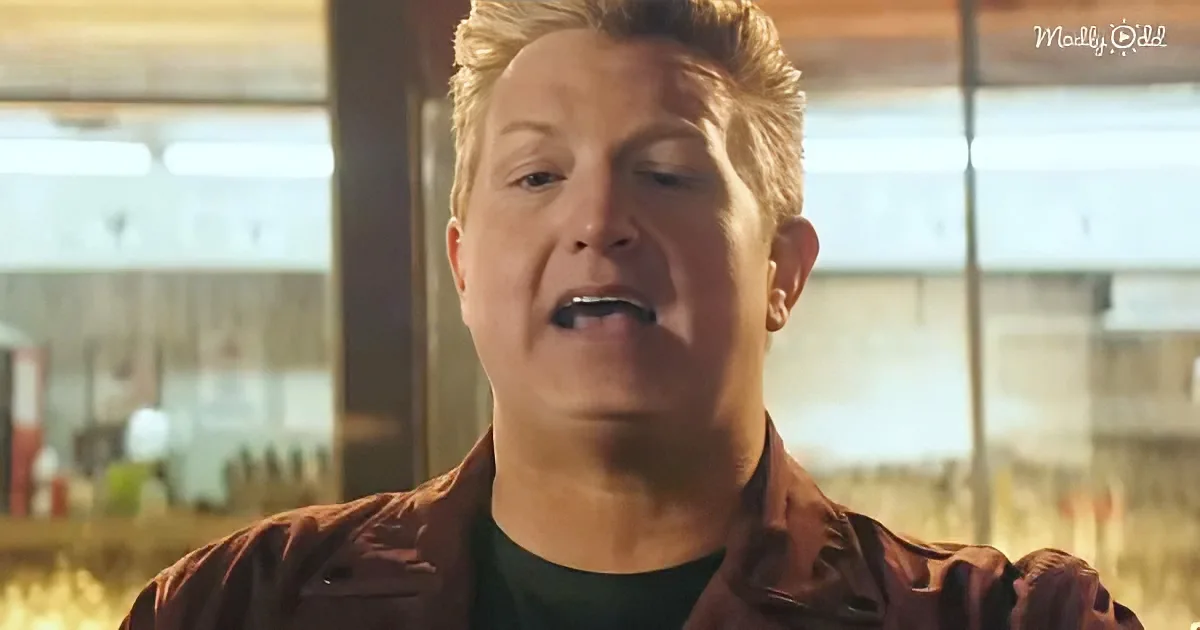 A man with short blonde hair, wearing a brown jacket, is speaking with his mouth open.