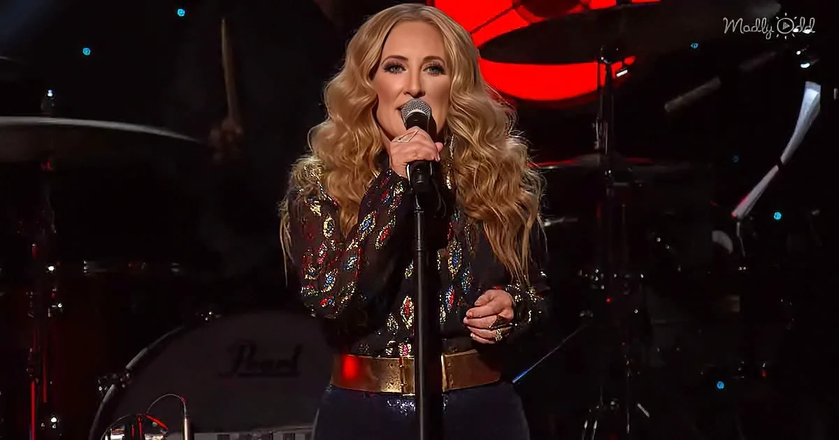 A woman with long blonde hair sings into a microphone on stage, wearing a black sequined top and gold belt.