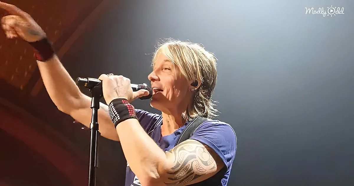 A person with long hair and a tattooed arm sings into a microphone on stage.