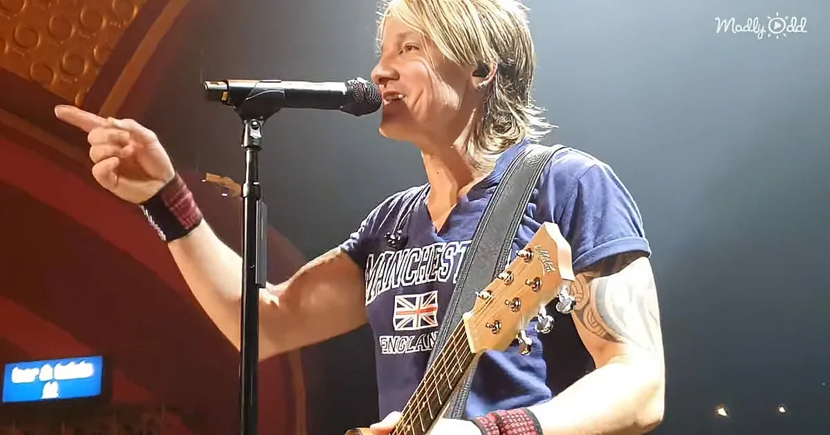 A person in a Manchester England shirt plays guitar and sings into a microphone, pointing to the left.