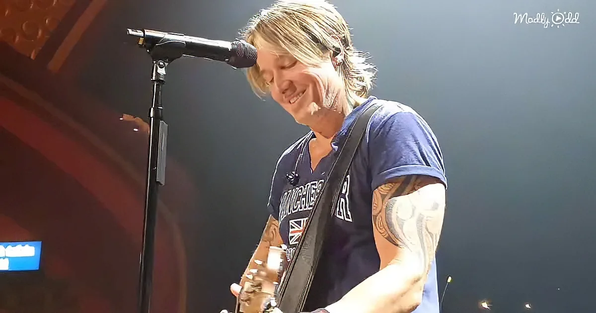 A smiling man with tattoos plays guitar on stage, wearing a blue shirt.