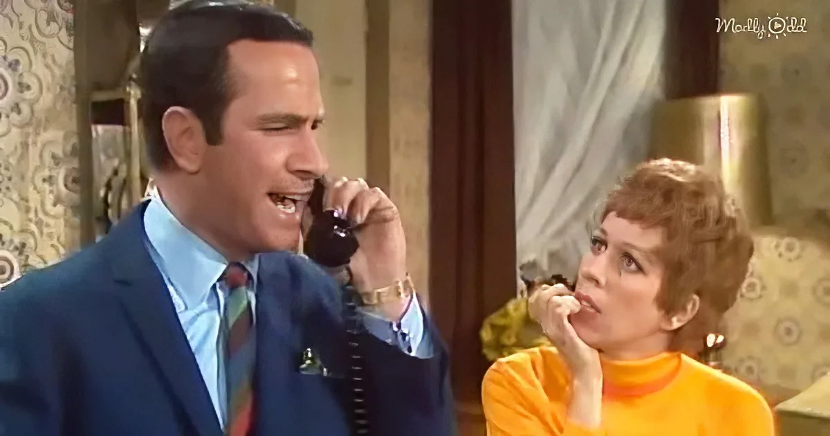 A man in a suit and tie is talking on a phone, while a woman in an orange top looks on with a hand on her chin.