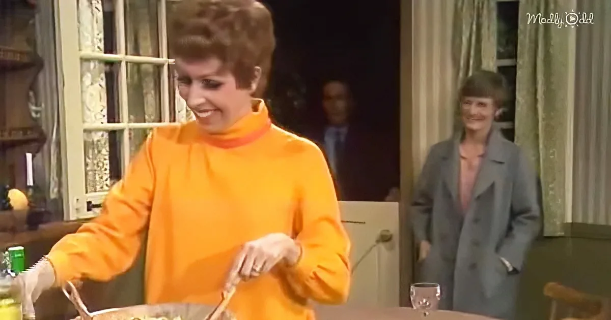 A woman in an orange sweater is cooking at a table, smiling, with another woman in a gray coat standing in the background.