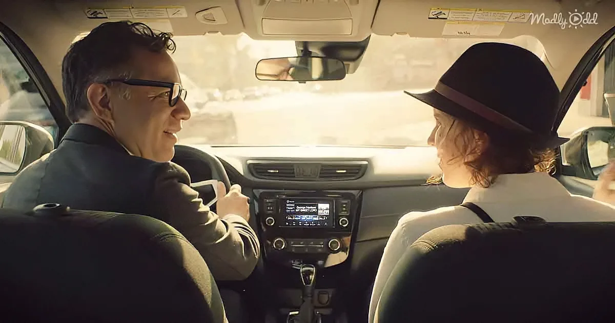 A man in a suit drives a car, smiling at a woman in a hat sitting in the passenger seat.