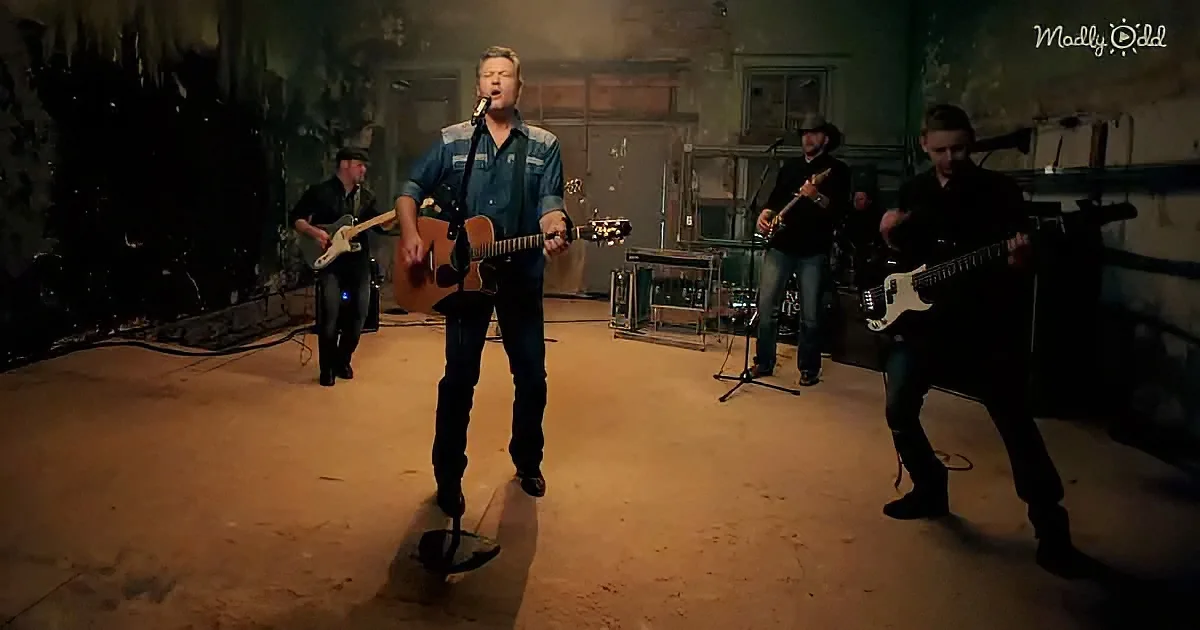 A man in a denim shirt sings and plays guitar in a dimly lit, rustic room with other musicians.