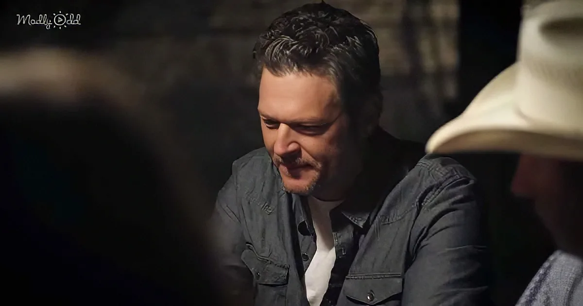 A man in a denim shirt and white shirt is smiling, looking down, with another person in a hat partially visible.