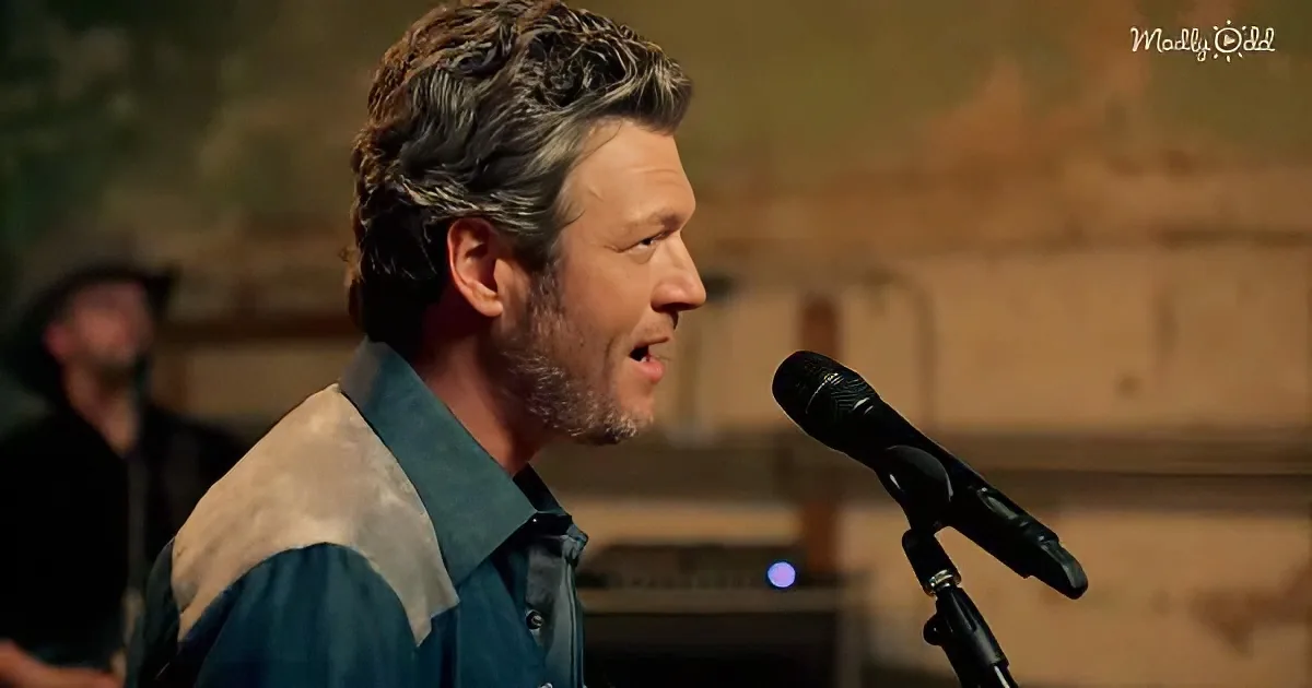 A man with short, curly hair sings into a microphone, wearing a blue and white shirt.