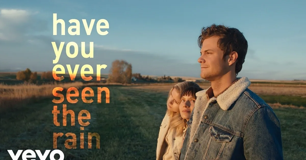 A man in a denim jacket stands in a field, looking off into the distance with two children behind him.