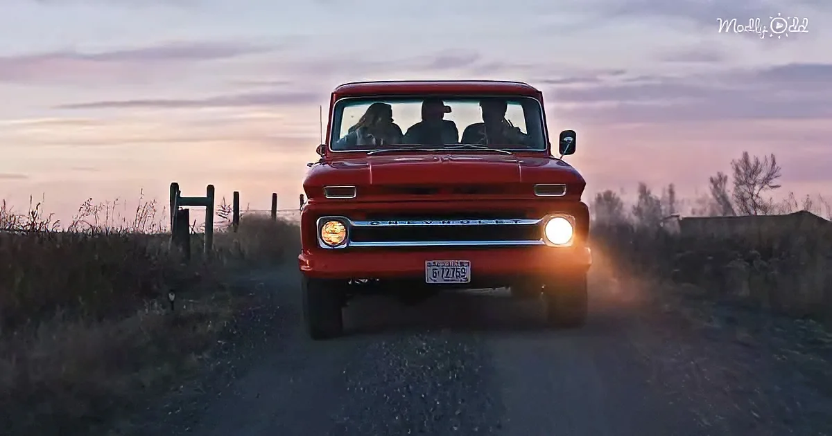 A red Chevrolet truck drives down a dirt road at dusk, with headlights on and three people inside.