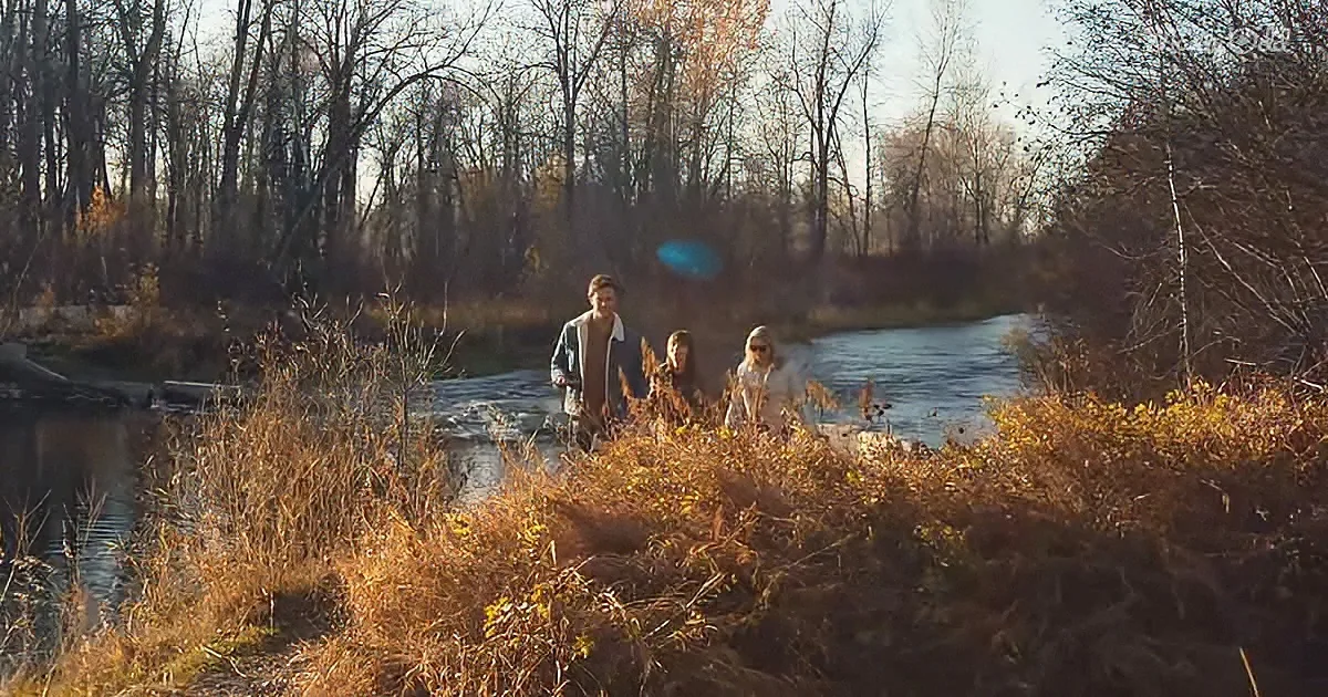 Three people walk through a shallow river surrounded by autumn foliage and trees.