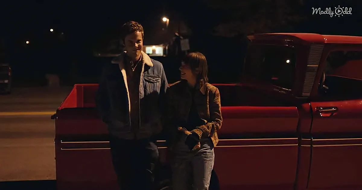 Two people stand beside a red vintage car at night, illuminated by streetlights.