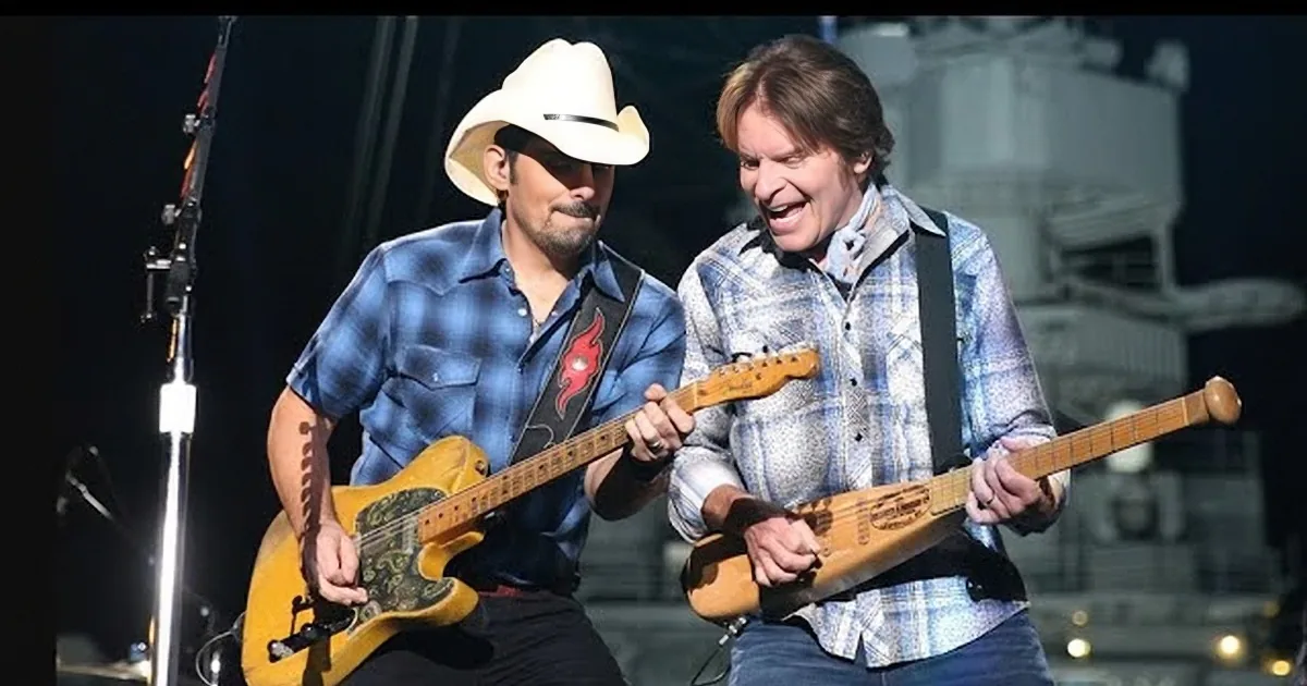 Two musicians perform on stage, one in a white hat, both playing guitars.