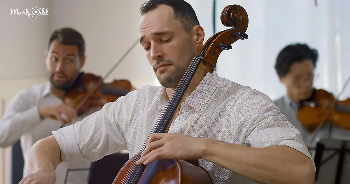 A man in a white shirt plays a cello with two violinists in the background.