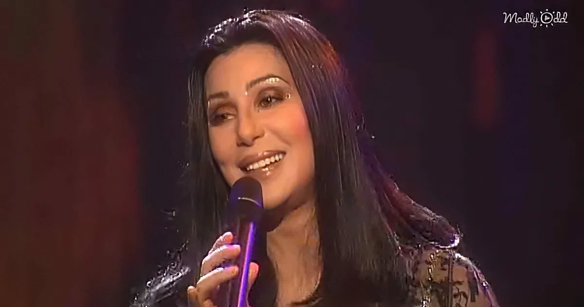 A woman with long dark hair smiles while holding a microphone, wearing a black outfit.
