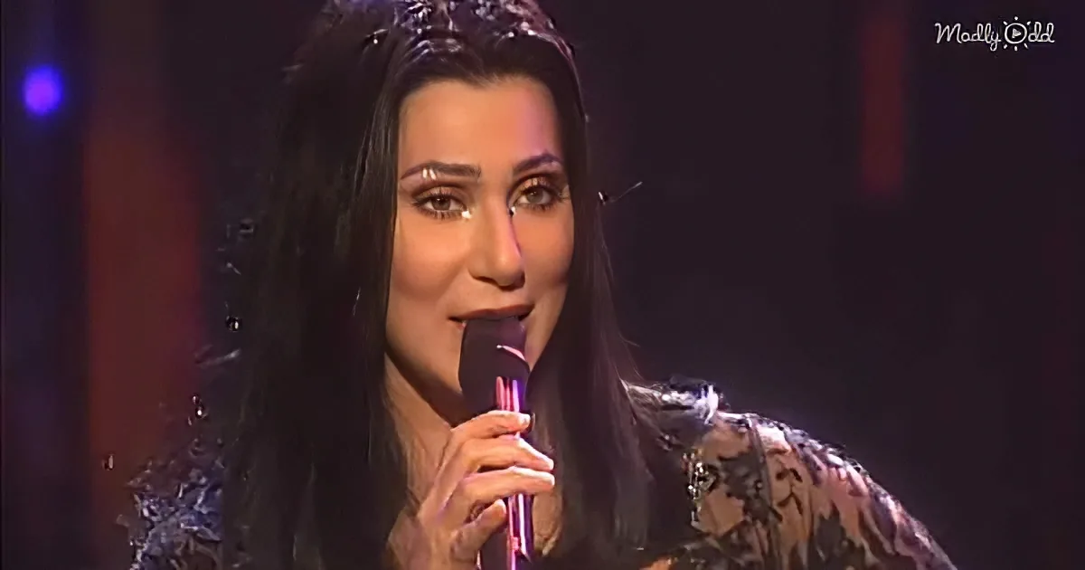 A woman with long dark hair and a tiara sings into a microphone, wearing a patterned top.