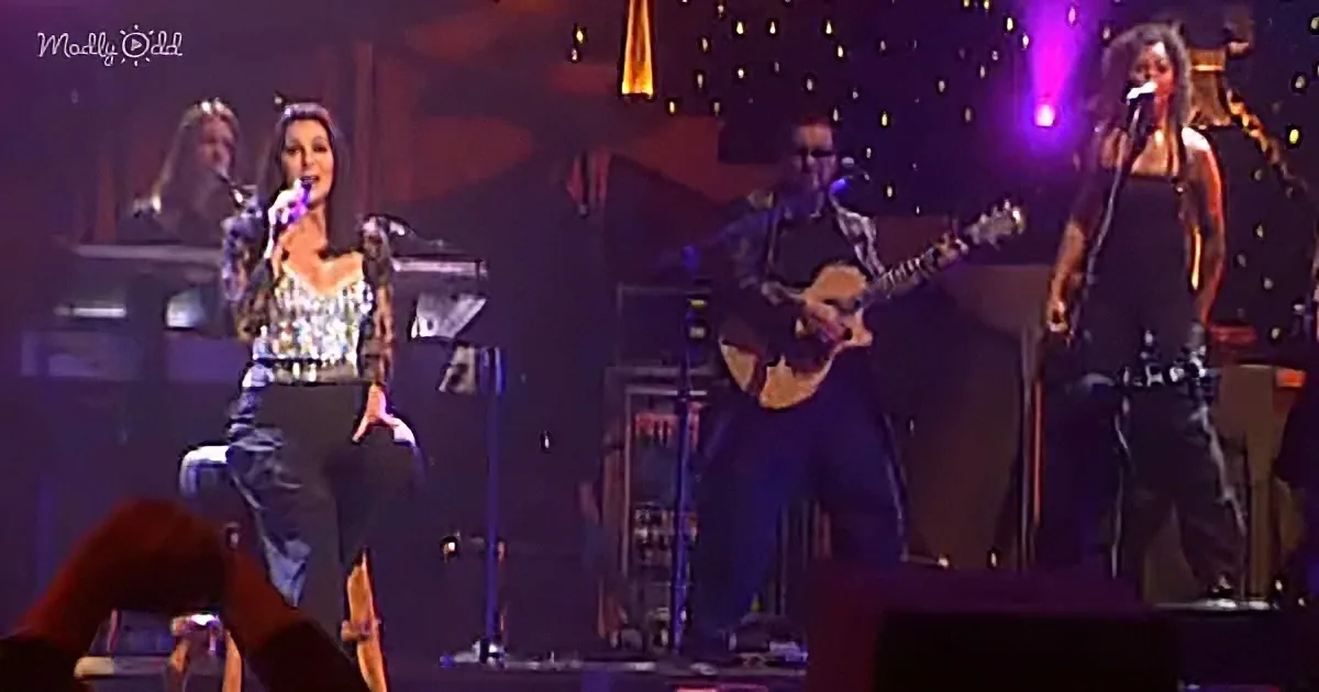 A woman in a sparkly top and black pants performs on stage with a guitarist and another woman singing.