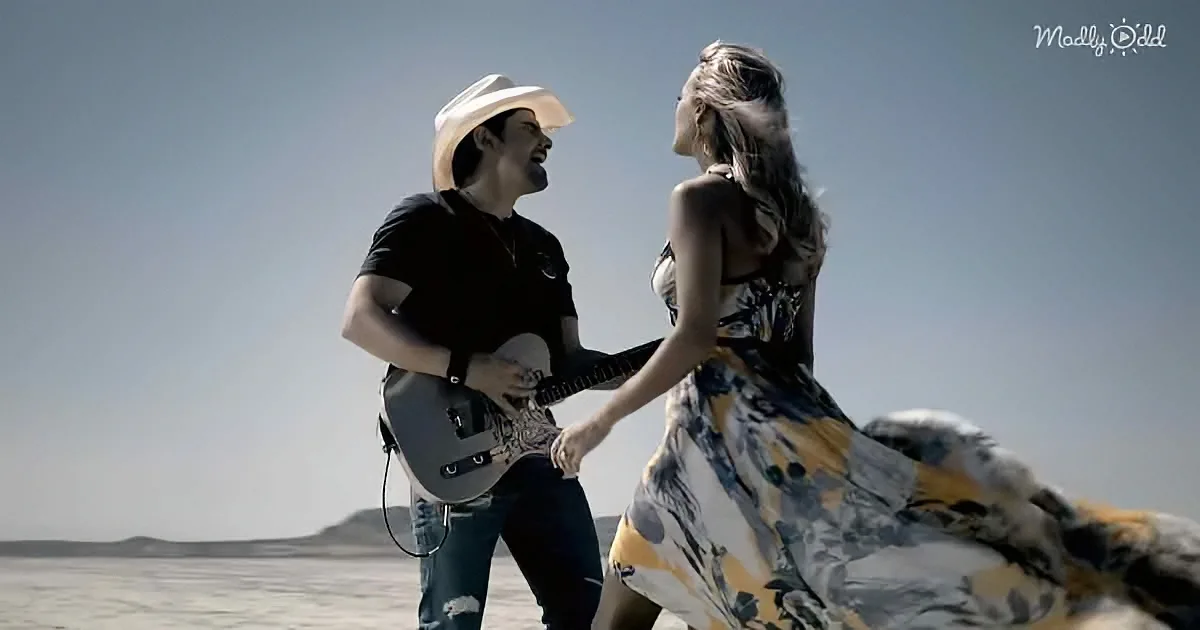 A man in a cowboy hat plays guitar while a woman in a floral dress dances beside him in a desert setting.