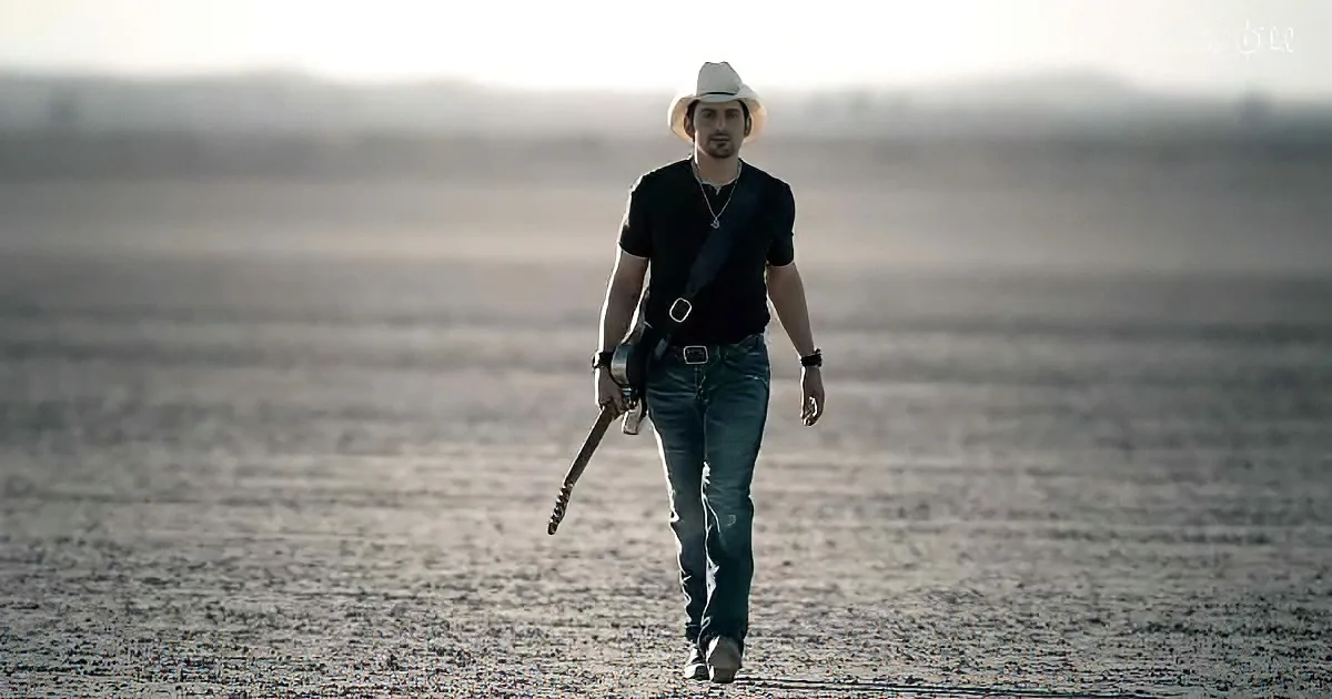 A man in a cowboy hat walks across a vast, barren landscape carrying a guitar.