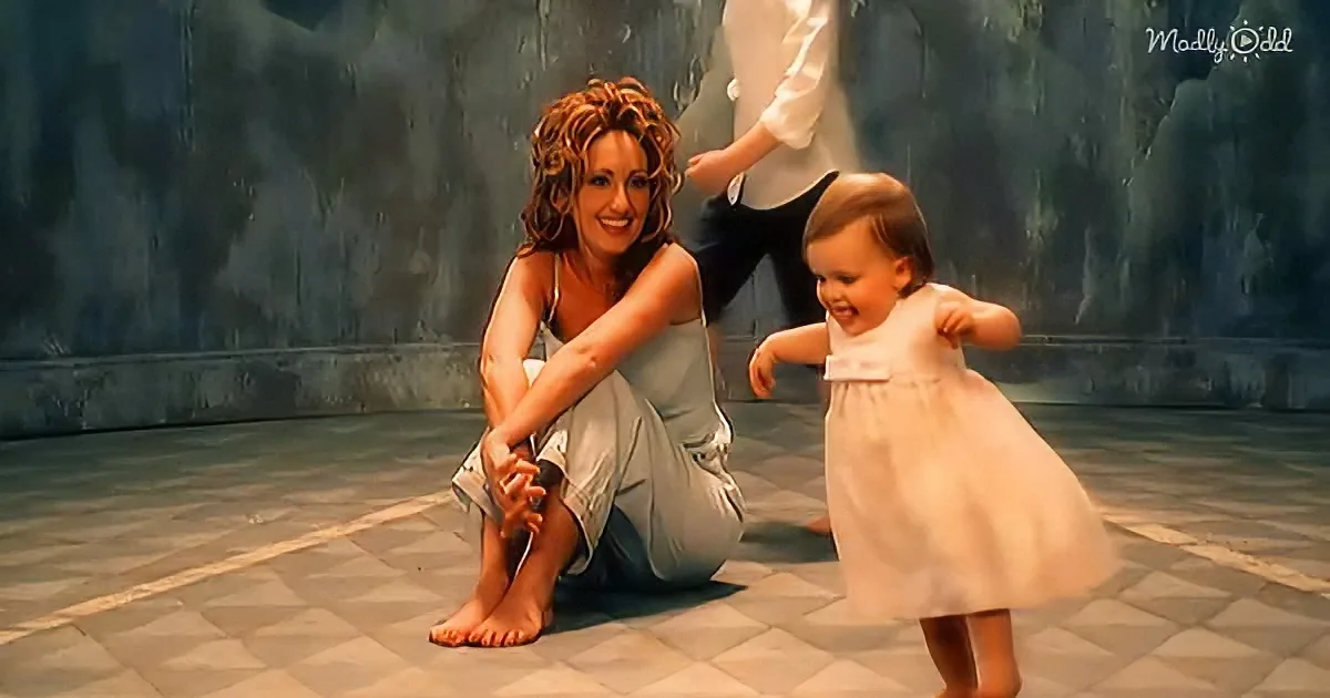 A woman in a white dress sits on the floor, smiling, with a baby in a white dress standing beside her.