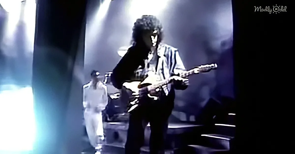 A guitarist in a striped shirt plays on stage under dramatic lighting, with two figures in the background.
