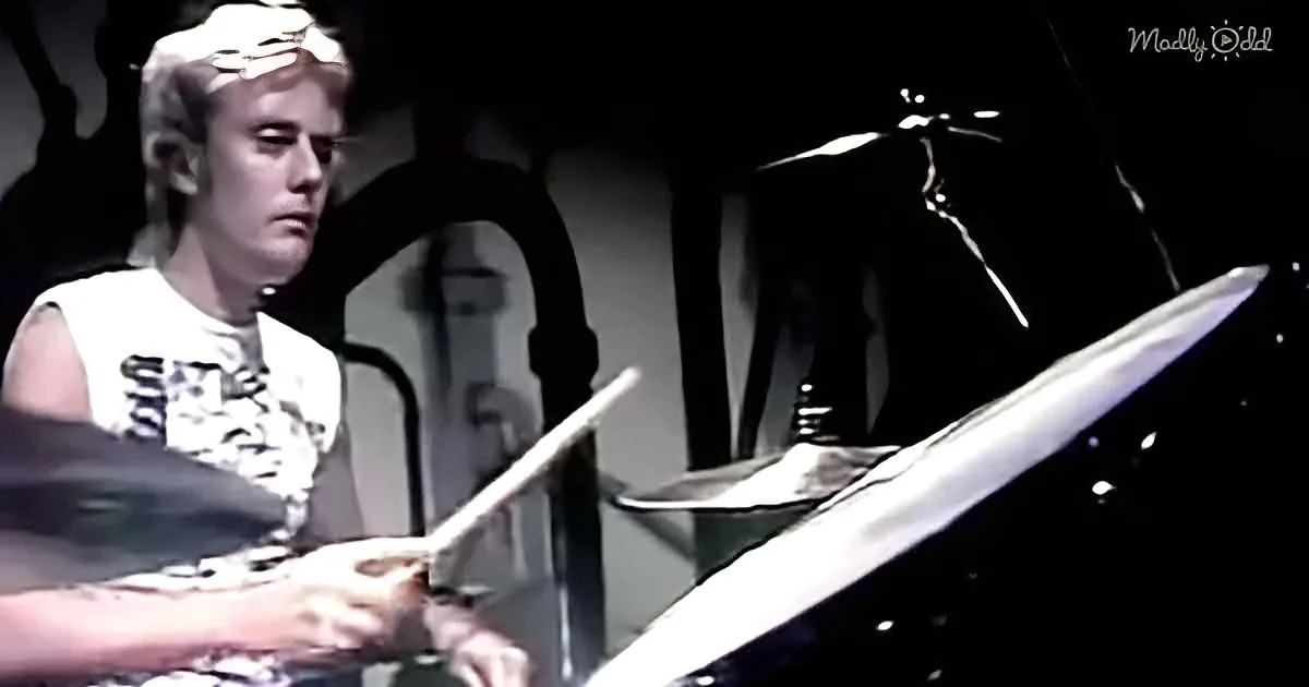 A person plays drums on stage, wearing a white shirt, with a microphone stand and drum set visible.