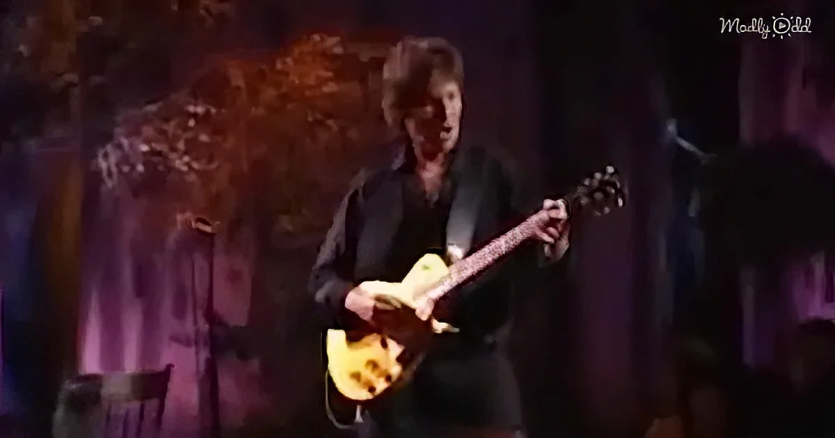 A person in a black suit plays a yellow electric guitar on stage, with a microphone and a crowd in the background.
