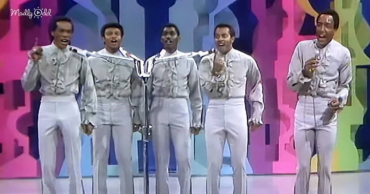 A group of five men in silver outfits perform on a colorful stage.