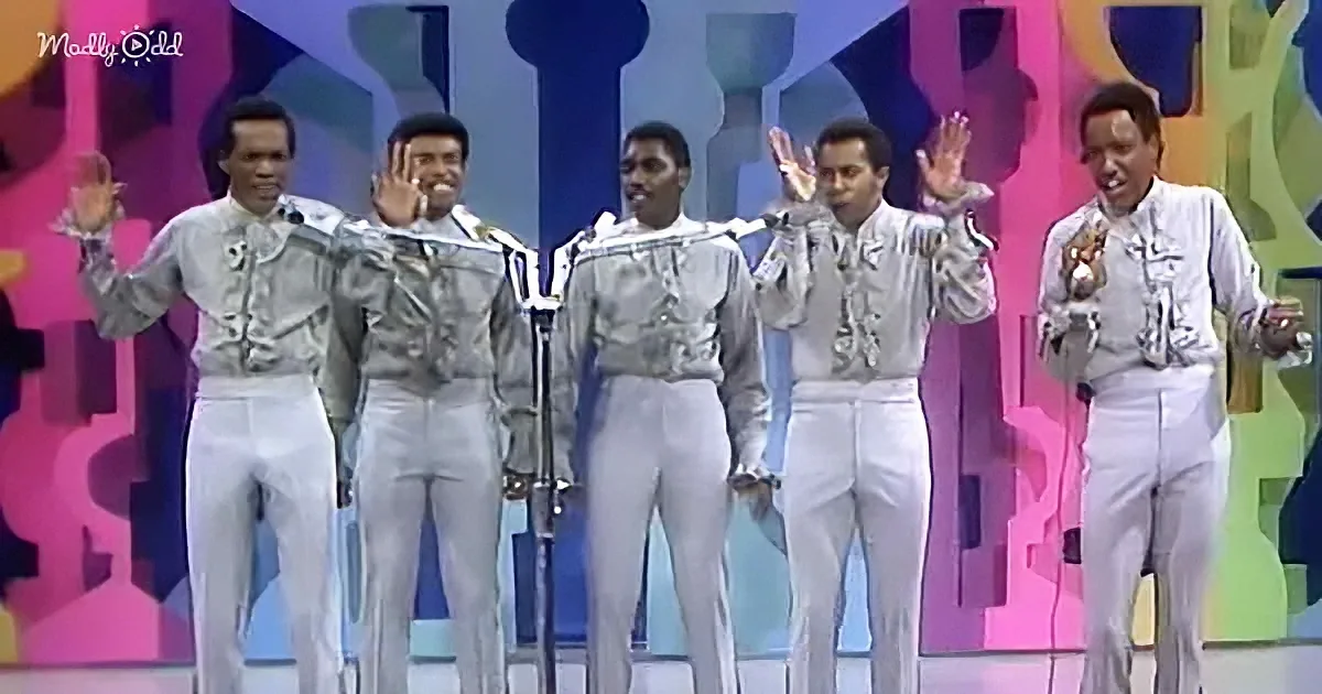 A group of five men in white outfits perform on stage with microphones and colorful background.