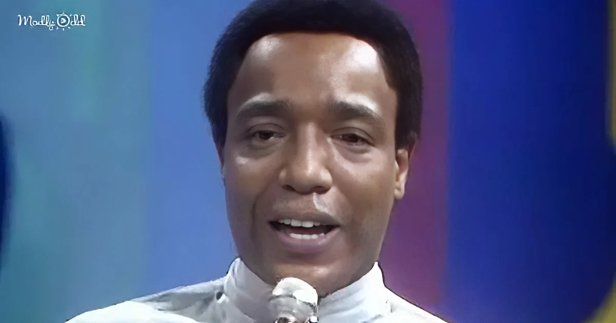 A man in a white shirt speaks into a microphone against a colorful background.