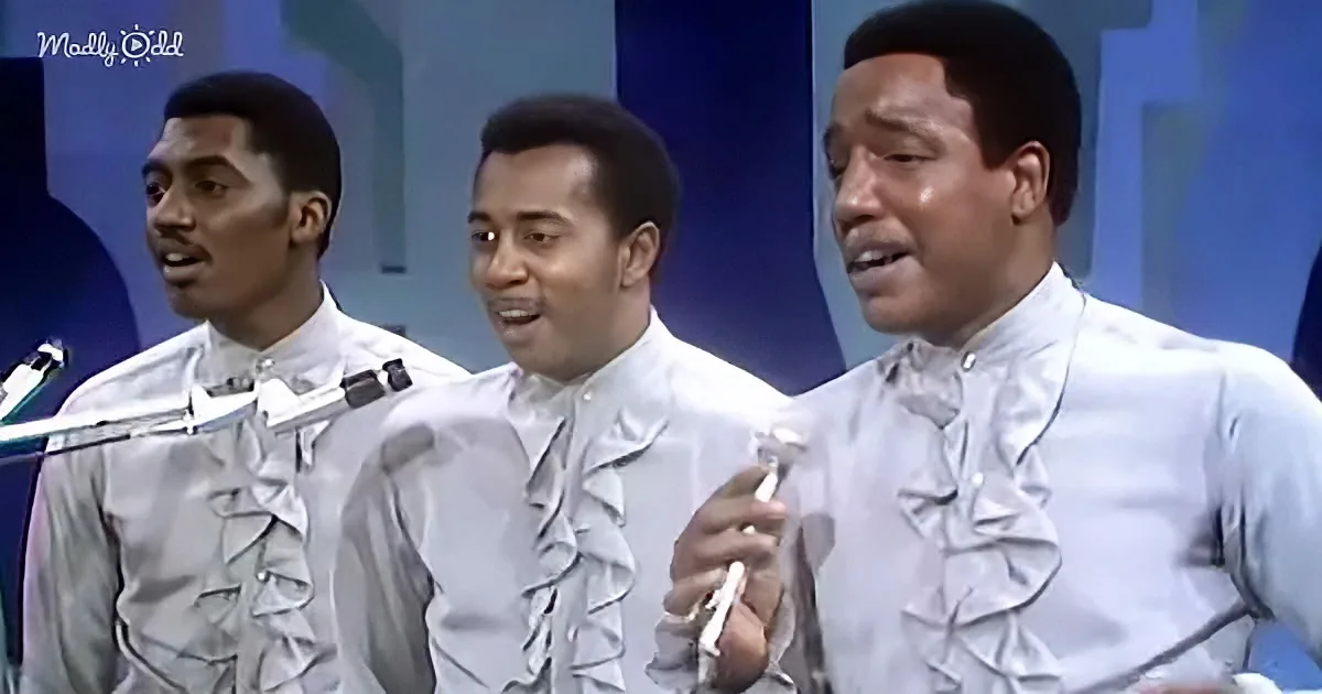 Three men in white ruffled shirts sing into microphones on a stage with a blue background.