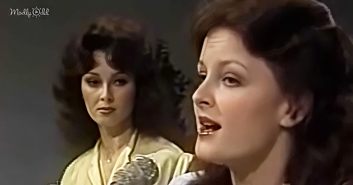 A woman with dark hair and a yellow top sings into a microphone, with another woman in the background.
