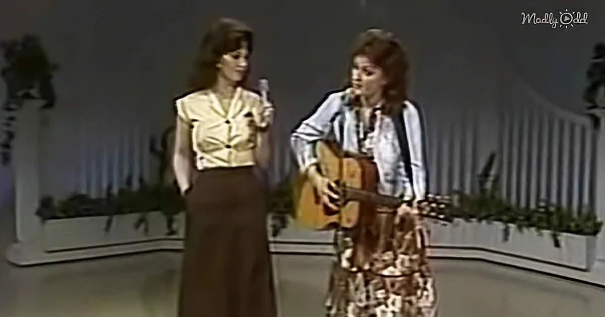 A woman in a floral dress plays guitar while another in a yellow top holds a microphone on stage.