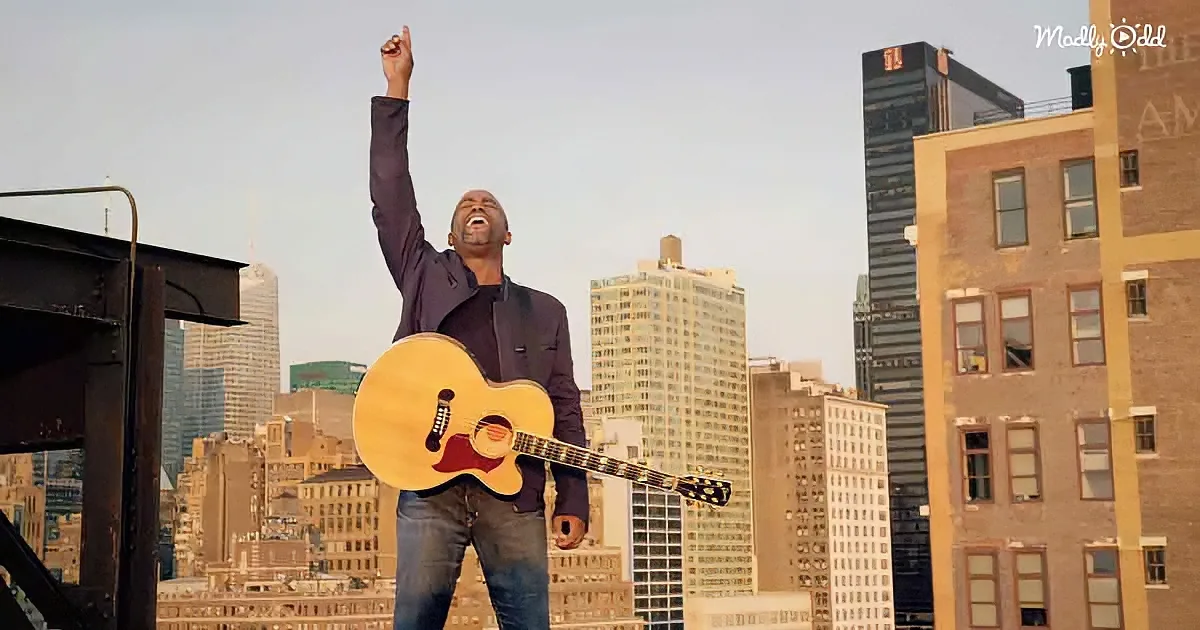 A man in a suit holds an acoustic guitar, raising his fist in the air against a cityscape backdrop.