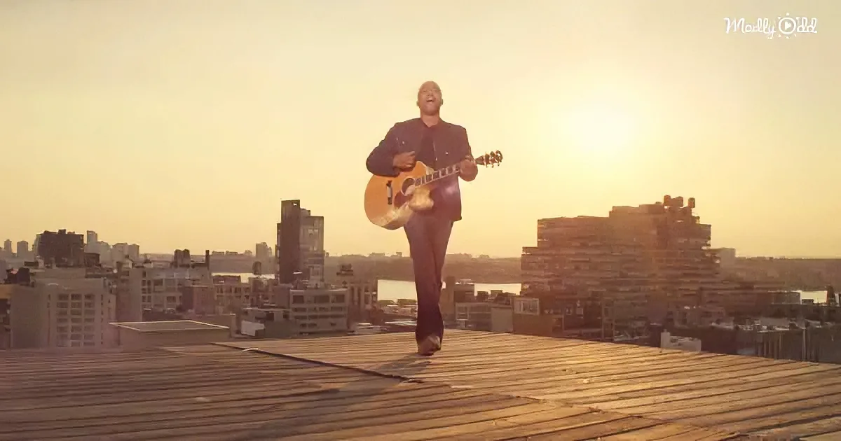 A man walks on a rooftop with a guitar, city skyline and sunset in the background.