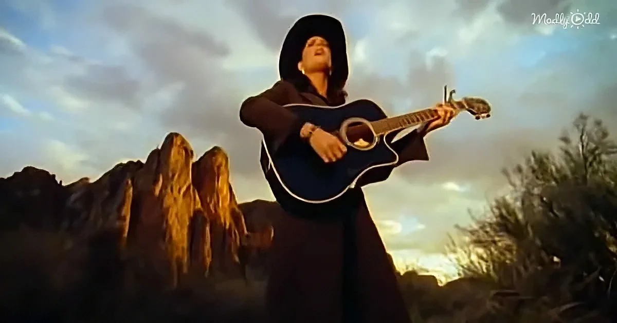 A person in a black outfit and cowboy hat plays a guitar against a desert backdrop with mountains.