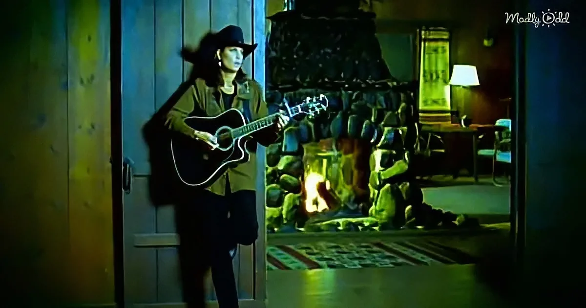 A person in a cowboy hat plays guitar near a lit fireplace in a rustic room.