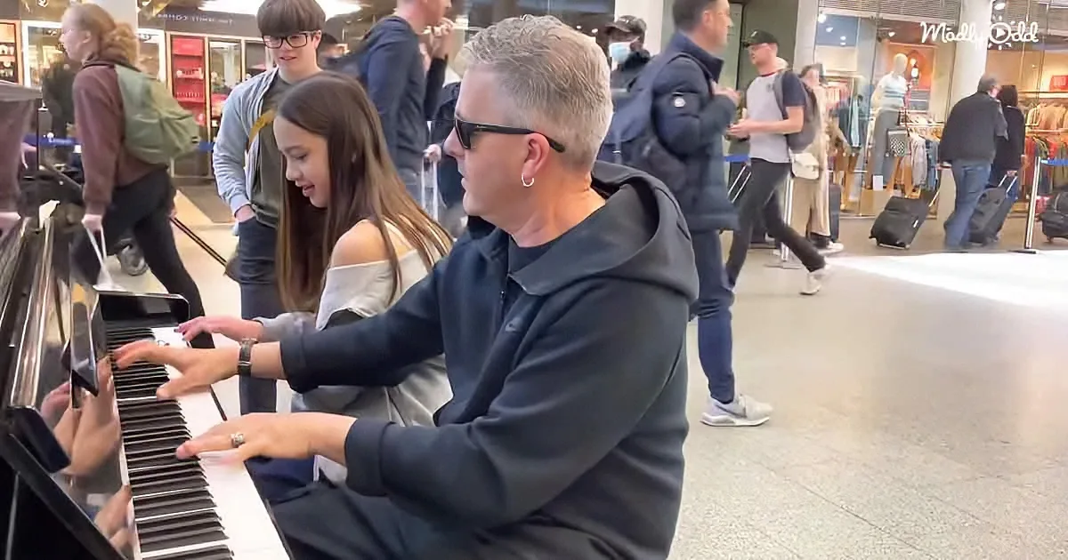 A man and a girl play piano together in a busy shopping area, surrounded by people walking by.