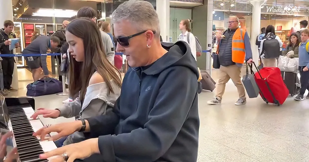 A man and a girl play a piano together in a busy airport terminal.