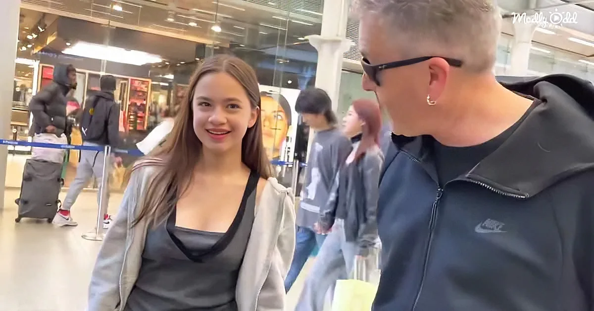 A woman in a grey jacket smiles while a man in a black Nike jacket looks on in a busy airport setting.