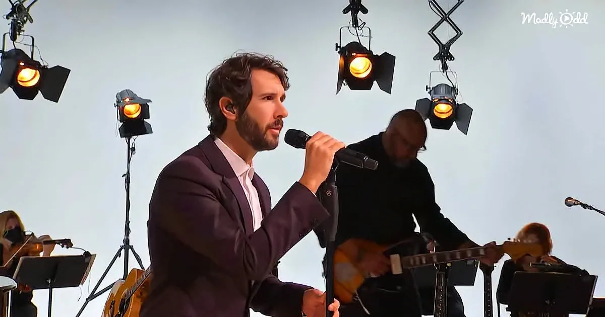 A man in a suit sings into a microphone on stage with musicians and spotlights.