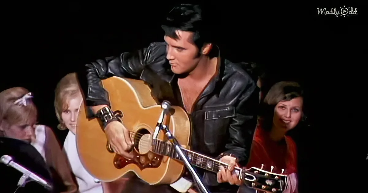 Elvis Presley in a black leather jacket plays an acoustic guitar on stage.