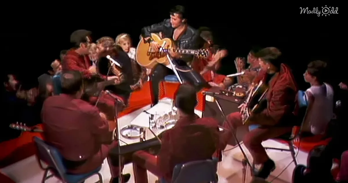 Elvis Presley plays guitar on stage surrounded by musicians and audience.