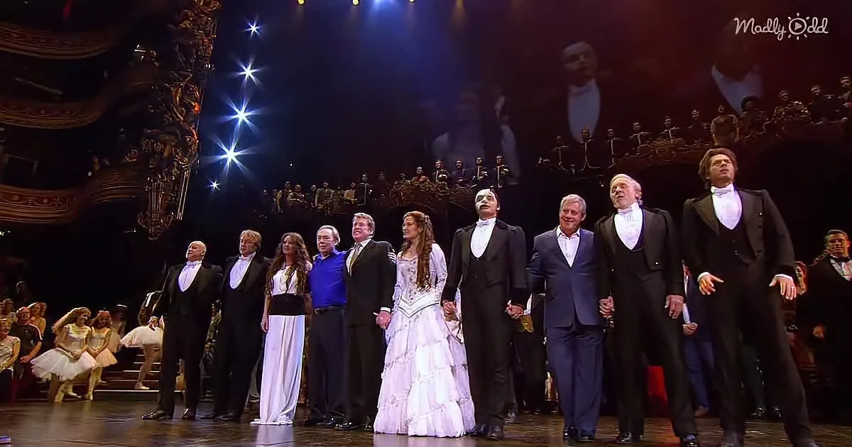 A group of performers in formal attire stands on stage, with dancers in the background, under dramatic lighting.