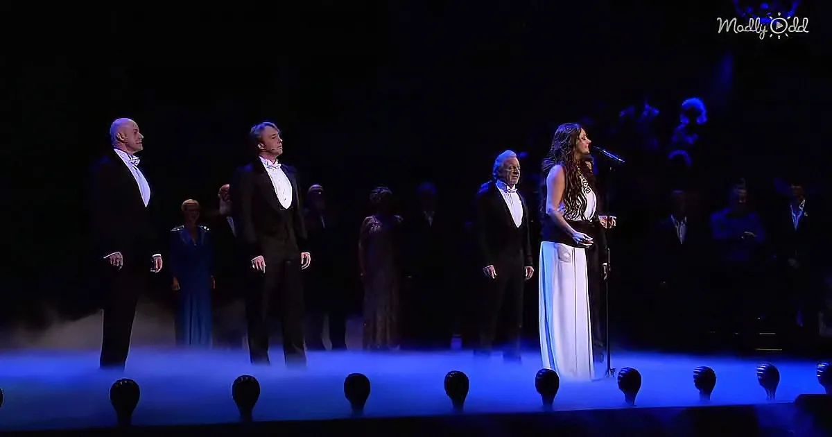 A woman in a white dress sings on stage with a man in a tuxedo, surrounded by other formally dressed performers.