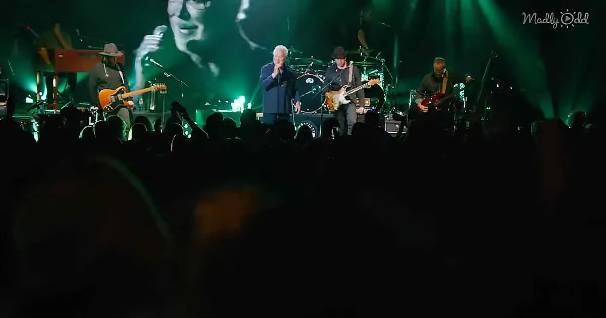 A band performs on stage with green lighting, audience in shadow, and a large screen showing a singer.