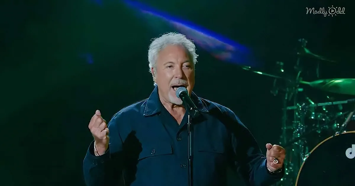 A man in a dark shirt sings into a microphone on stage, with a drum set and green lighting in the background.