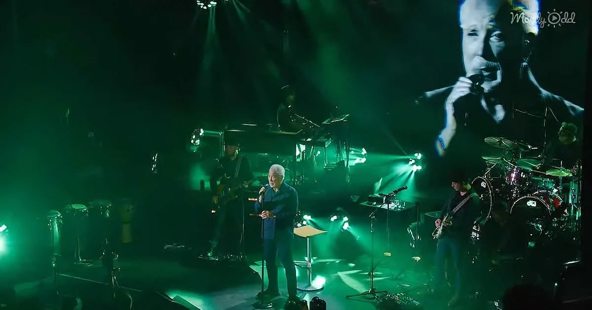 A man sings on stage with a band, illuminated by green lights, with a large screen showing a close-up of him.